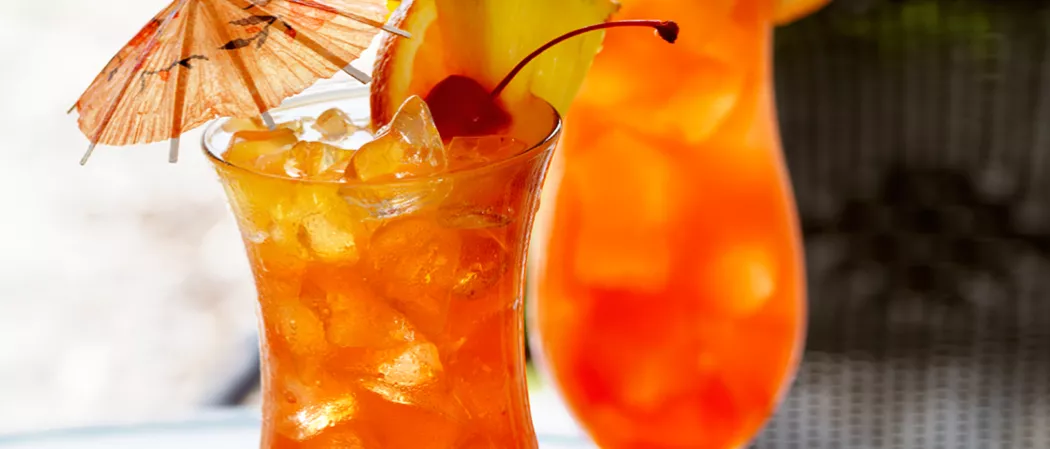 A glass of Watermelon Mai Tai, garnished with ice cubes, a cherry, orange and  pineapple slice on the rim of the glass, and a cocktail umbrella