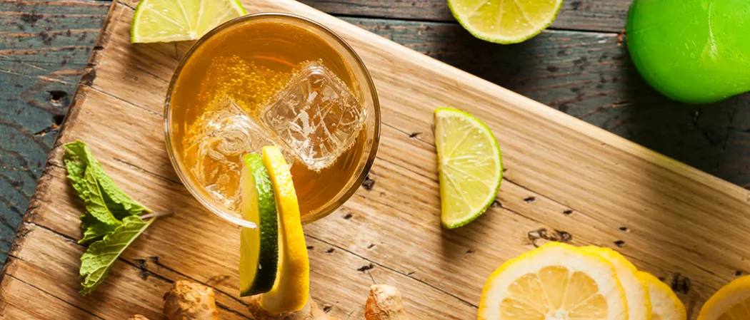 A glass of Mexican Mule, garnished with ice cubes and wedges of lime and lemon on the rim, alongside slices of lime and mint leaves on a wooden tray