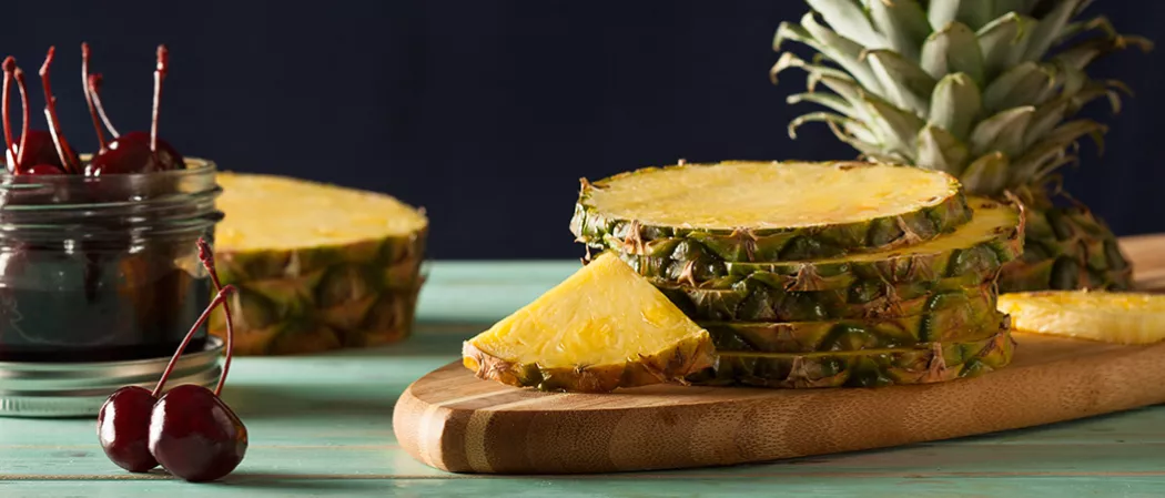 Four round slices of pineapple arranged on a wooden tray. along with  a jar of cherries.