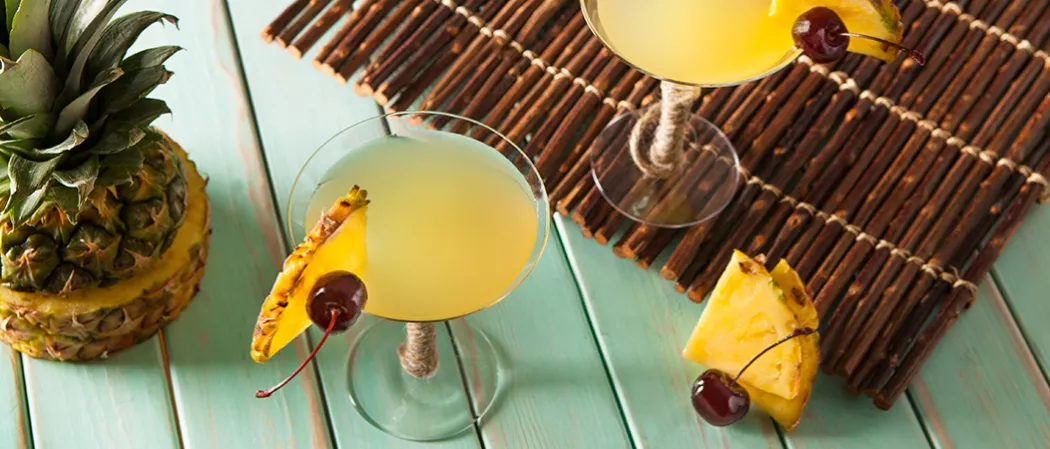 Two glasses of Pina Colada Martini, each garnished with a slice of pineapple and a cherry on the rim, one placed on a wooden mat. Alongside a half-cut pineapple, cherry, and slices of pineapple.