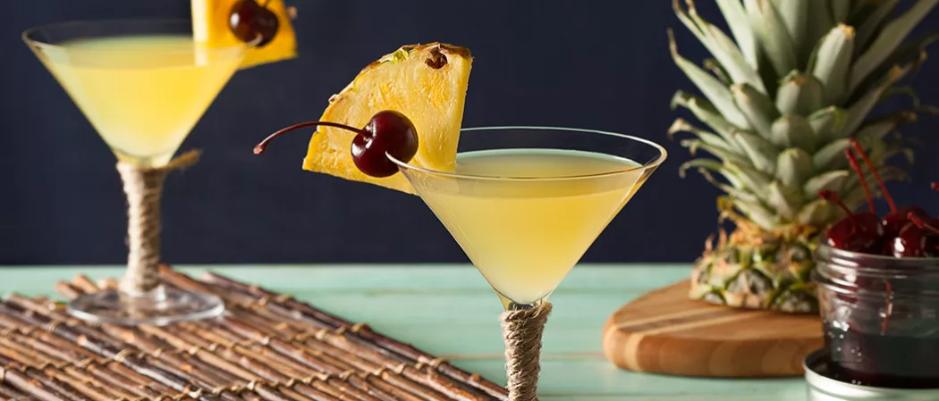 Two glasses of Pina Colada Martini, each garnished with a slice of pineapple and a cherry on the rim, placed on a wooden mat.