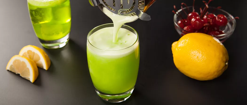 Two glasses of Fresh Midori® Sour. a person is pouring the drink using a cocktail strainer in a glass. Alongside a bowl of cherries, a lemon, and lemon wedges.