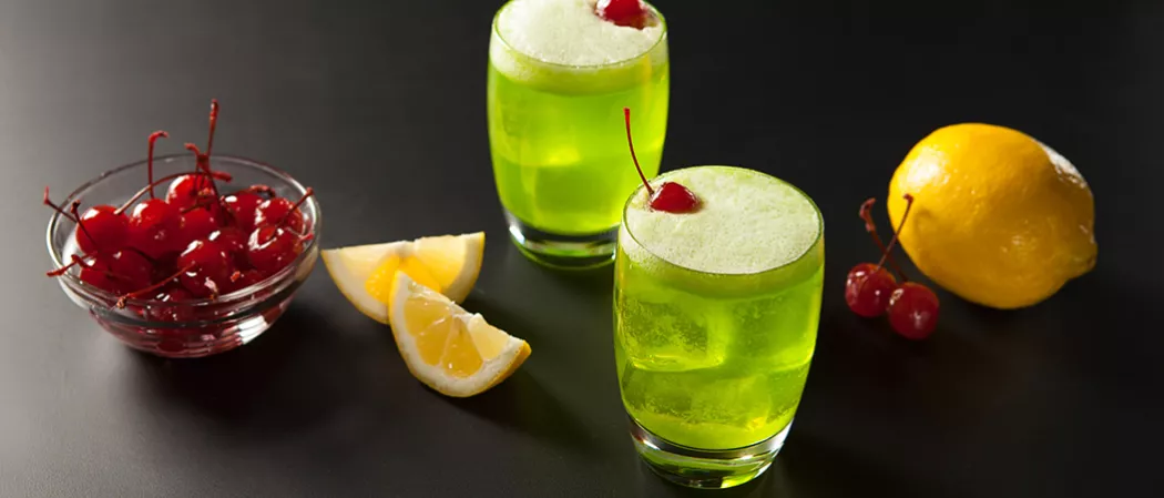Two glasses of Fresh Midori® Sour garnished with a cherry, alongside two additional cherries, a bowl full of cherries, a lemon, and lemon wedges.