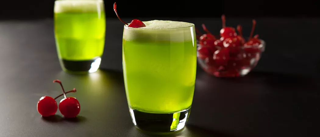 Two glasses of Fresh Midori® Sour garnished with a cherry, alongside two additional cherries and a bowl full of cherries.