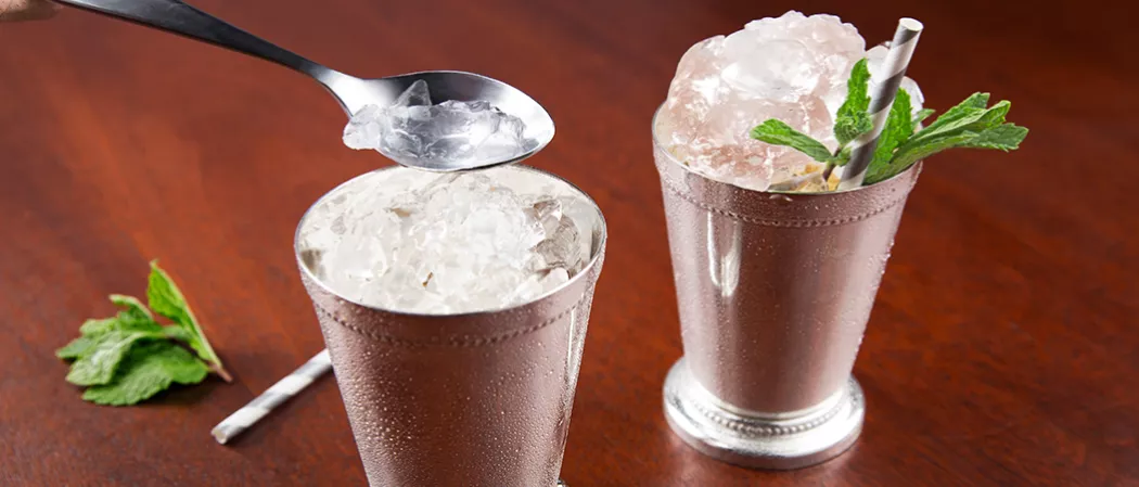 Two silver glasses of Maker's Mark Mint Julep, one with a straws and crushed ice, garnished with mint leaves. In the second glass, a person is filling crushed ice using a spoon alongside with a straws and mint leaves.