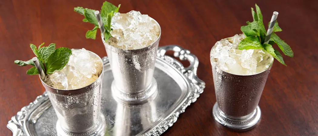 Three silver glasses of Maker's Mark Mint Julep with a straws and crushed ice, garnished with mint leaves. Two glasses are on a silver tray, and one is on the table.