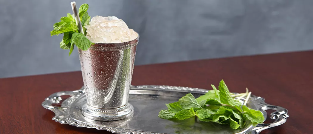 Silver glass of Maker's Mark Mint Julep with a straws and crushed ice, garnished with mint leaves, alongside some additional mint leaves on a silver tray.