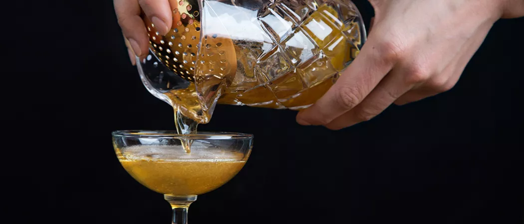 A person is pouring the Bellini Manhattan from a jar using a cocktail strainer into a glass.