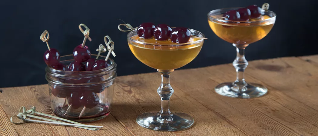 Two glasses of Bellini Manhattan garnished with cherries on a toothpick, alongside a cocktail strainer and a jar half filled with Bellini Manhattan.