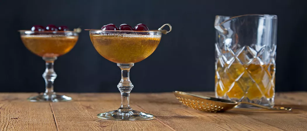 Two glasses of Bellini Manhattan garnished with cherries on a toothpick, alongside a cocktail strainer and a jar half filled with Bellini Manhattan.