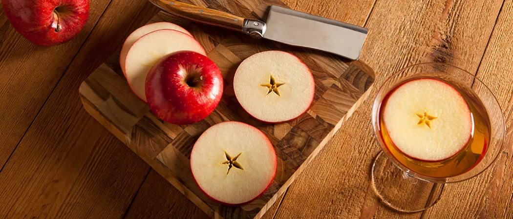Sliced apple and a knife on a chopping board next to a glass of Apple Manhattan garnished with an apple slice.