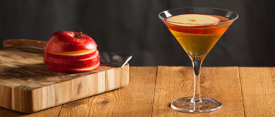 Glass of Apple Manhattan garnished with an apple slice, alongside sliced apple and a knife on a chopping board
