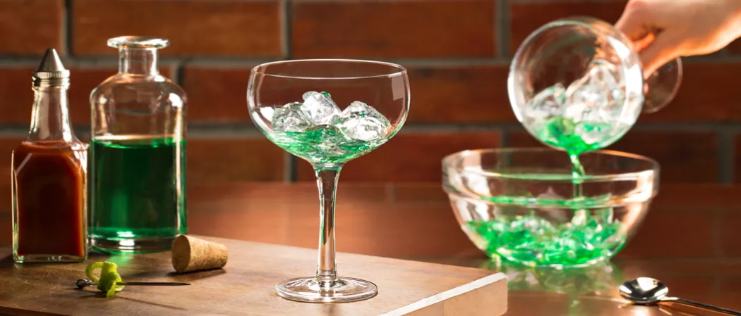 Glass with a small amount of Absinthe and crushed ice, next to a bottle of Absinthe, bitters, a lime twist, and a bottle stopper