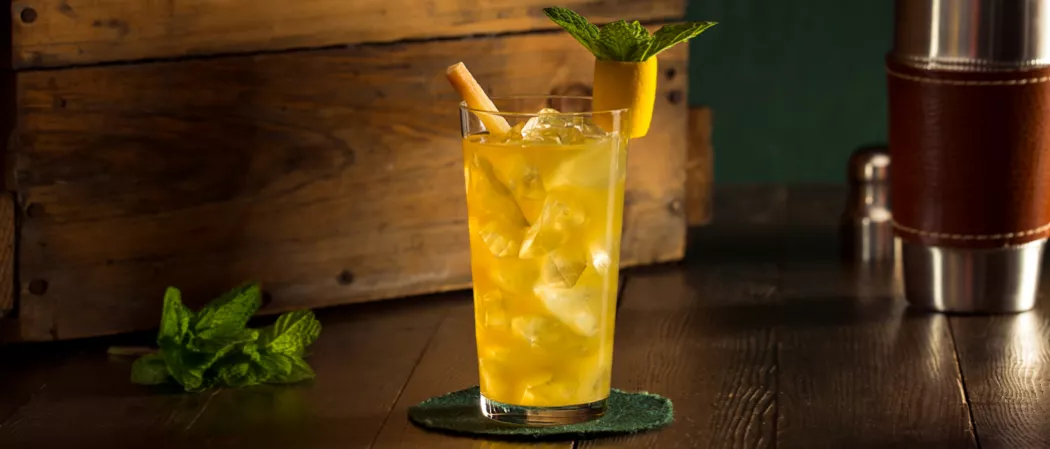 Glass of David Beam's 1838 Cocktail on a coaster, garnished with a sugar cane stir stick and a pineapple parrot, along with mint leaves and a shaker.