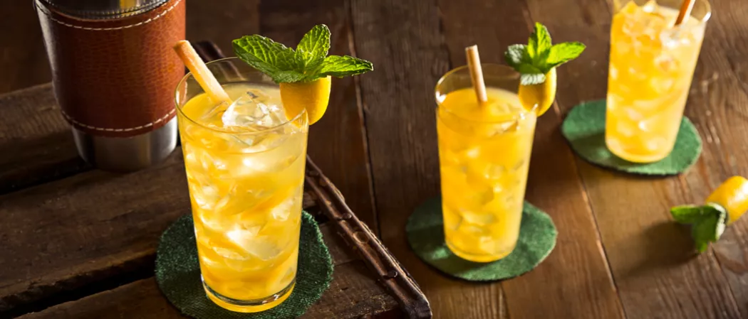 Three glasses of David Beam's 1838 Cocktail, one on a tray with a shaker, two on the table, each adorned with a sugarcane stir stick and pineapple parrot