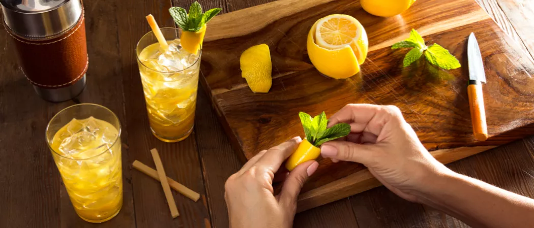 A person is creating a pineapple parrot while two glasses of David Beam's 1838 Cocktail, one garnished with a sugarcane stir stick and pineapple parrot, sit next to a shaker