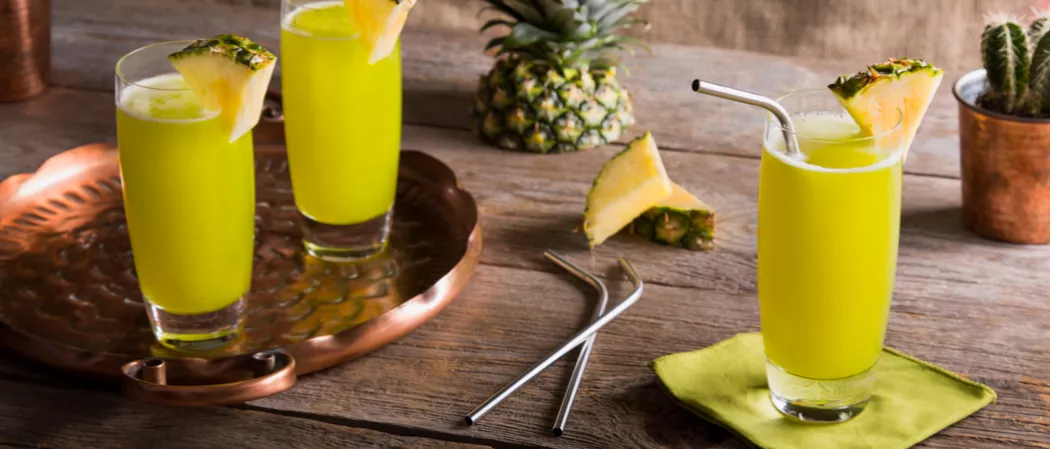 Three glasses of Midori® Cactus Juice, two glasses garnished with pineapple wedges on a brass tray, and the third glass garnished with a pineapple wedge on a green napkin with a straw. 
