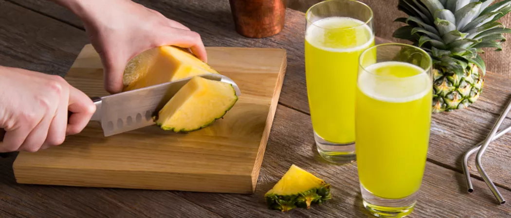 A person is cutting pineapple wedges on a chopping board, alongside two glasses of Midori® Cactus Juice, half a pineapple, straws, and a pineapple wedge.