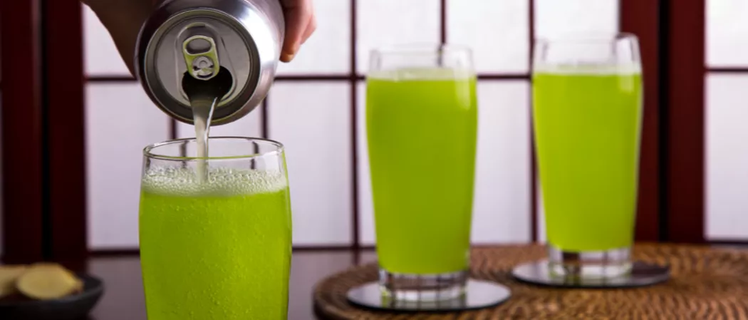 a person pouring green midori and ginger beer  in a glass, alongside two  glass of  same drink on a table