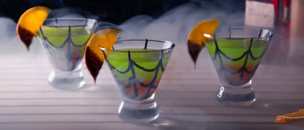 Three glasses of Spider's Kiss with a chocolate web drawn, garnished with chocolate-dipped orange slices and smoke spreading around.
