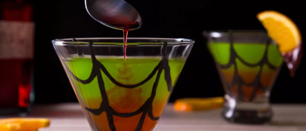 A person is pouring drizzled grenadine into a glass of Spider's Kiss with a chocolate web drawn.