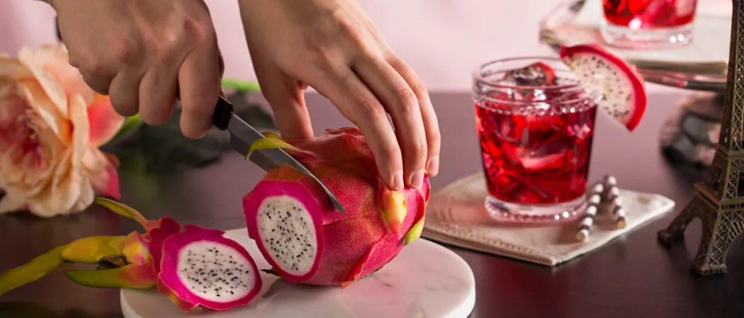 Person slicing dragon fruit with knife alongside a glass of Paris Dream garnished with slice of dragon fruit