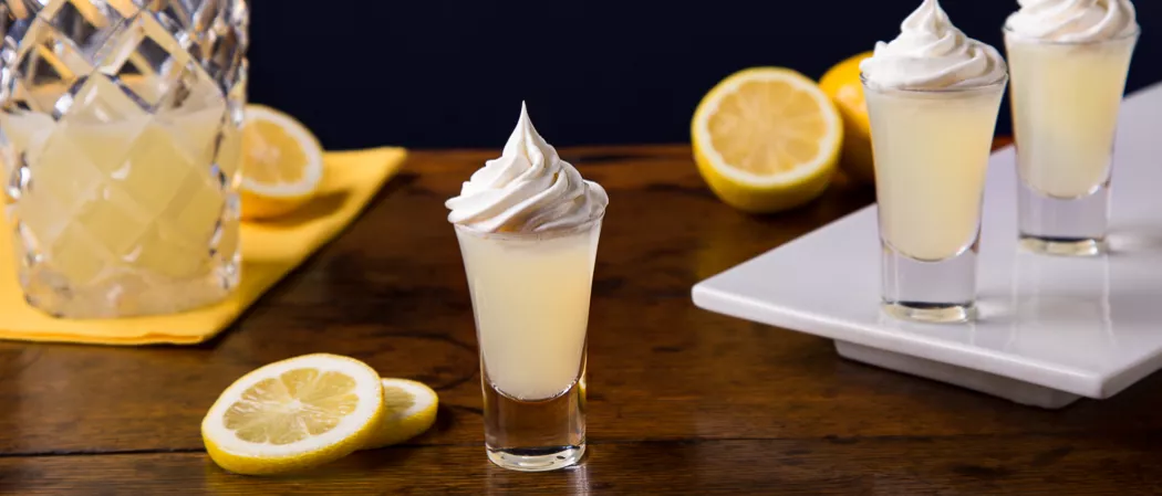 A glass of Ooh La La Lemon, topped with whipped cream, is accompanied by lemon slices. two glasses of same drink  sits on a tray, alongside few lemons. Additionally, a jar  on the paper mat with halved lemons.