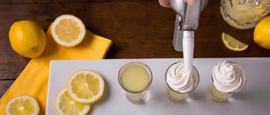 Three glasses of Ooh La La Lemon, one topped with whipped cream, another being garnished by a person, accompanied by lemon slices, all on a white tray, over a yellow paper cloth. 