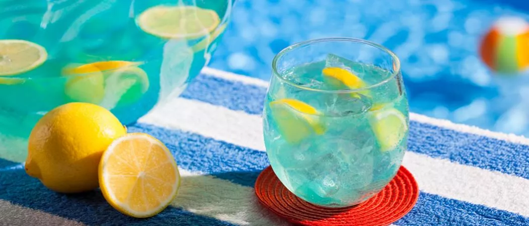 A glass of ice-cold drink with lemon slices, alongside a lemon and a summer fishbowl of same drink.