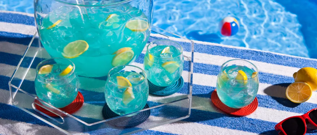 A glass of Pool Party Punch with ice and lemon slices on red coaster alongside three glasses of same one on red coaster and a jar of Pool Party Punch on transparent tray