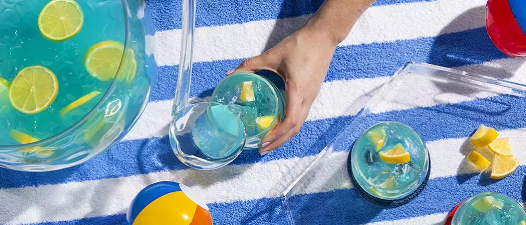 A person pouring a lemon-infused ice-cold drink from a  summer fishbowl into a glass alongside a glass of same drink.A tray with two glasses and a summer fish bowl containing a Ice-cold drink garnished with lemon.