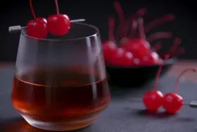 A glass of dark-colored cocktail garnished with three maraschino cherries on a metal pick. In the background, a black bowl filled with additional maraschino cherries.